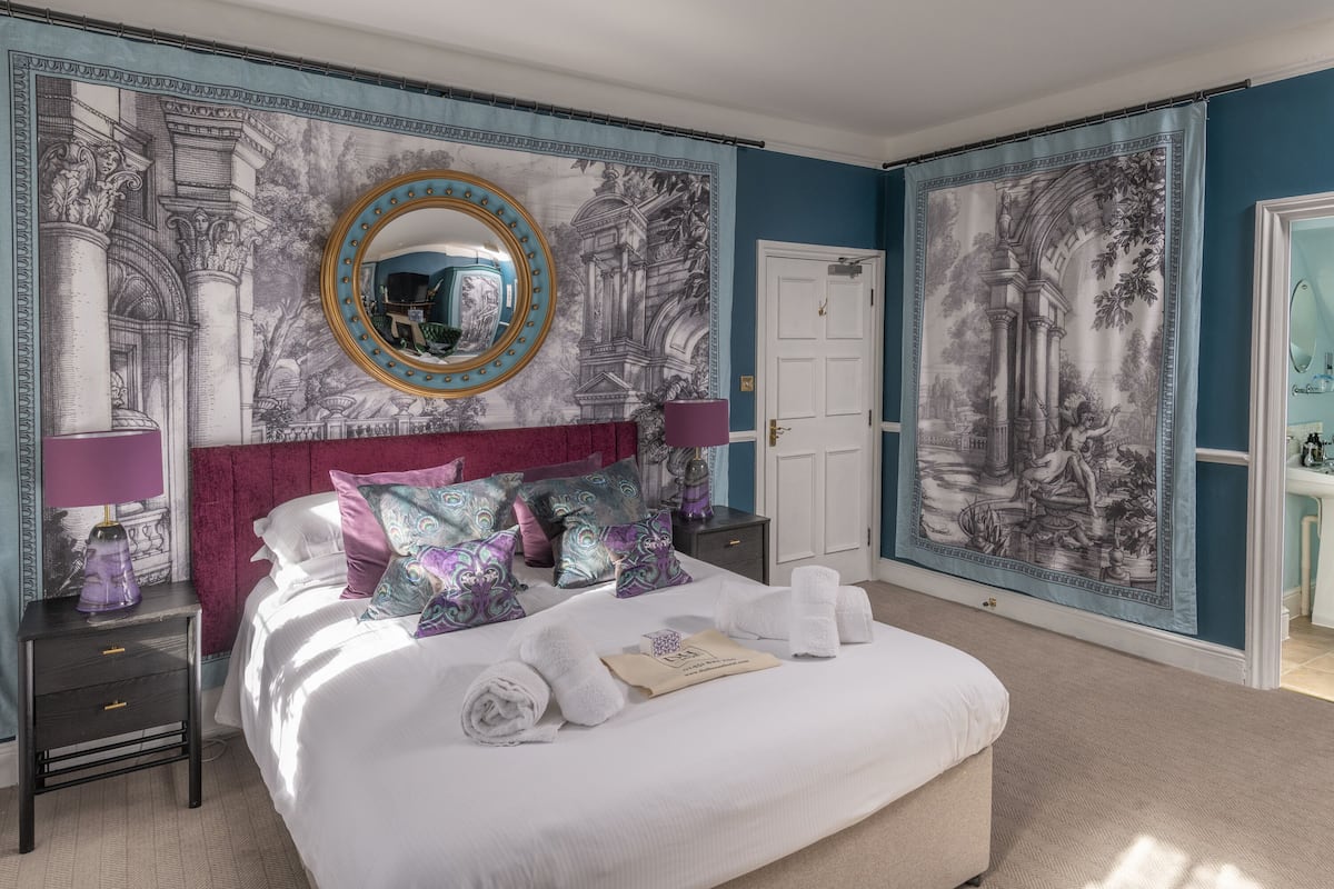 Quarto com uma cama grande, cabeceira, mesa de cabeceira, abajur, espelho e tapeçaria na parede.