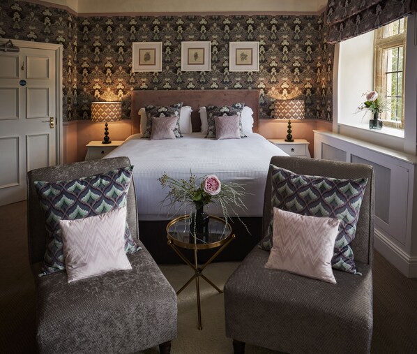 Une chambre à coucher avec un grand lit, deux fauteuils, une petite table avec un vase, un papier peint à motifs et des tableaux encadrés accrochés au mur.