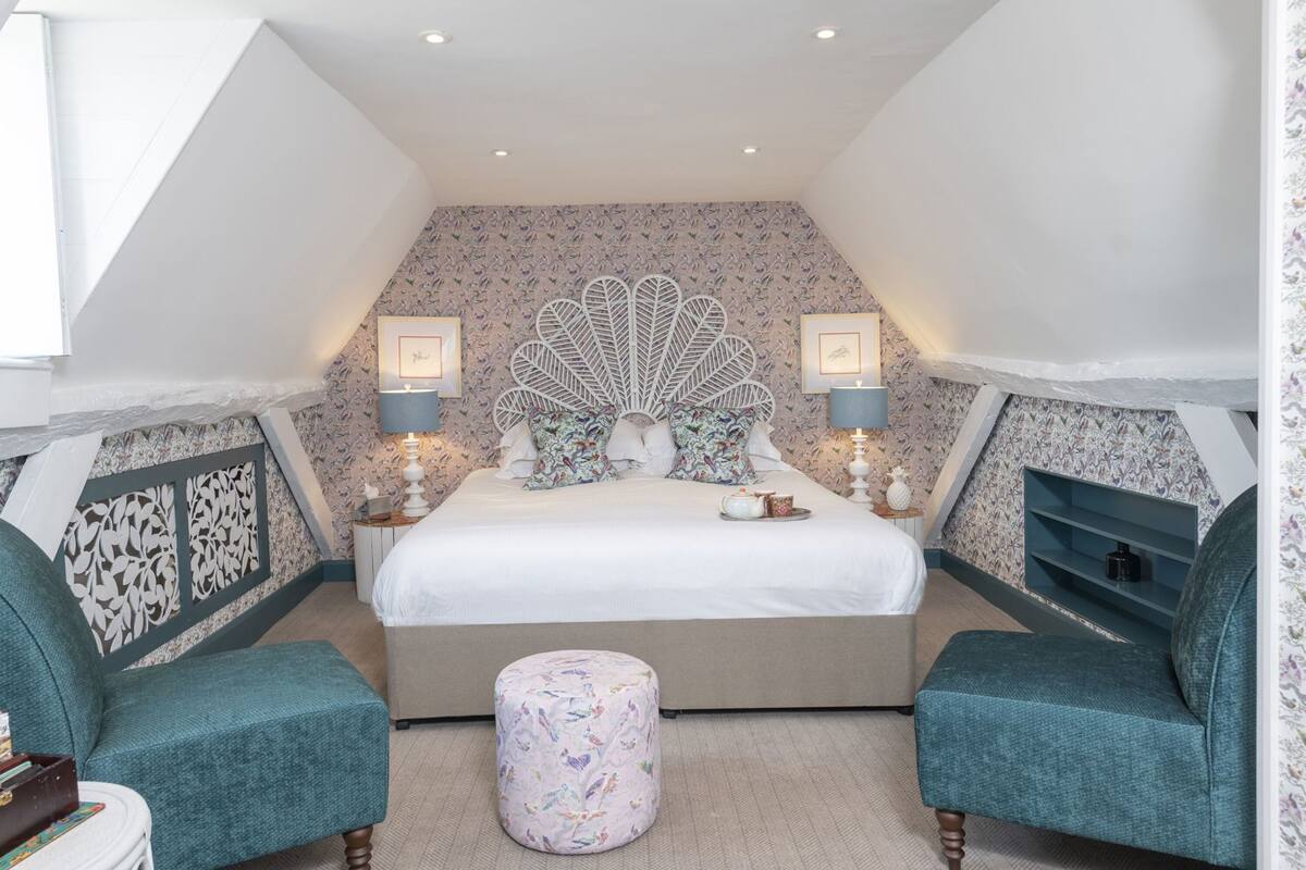 Quarto com cama grande, duas mesas de cabeceira, cabeceira decorativa, lareira e duas poltronas combinando.