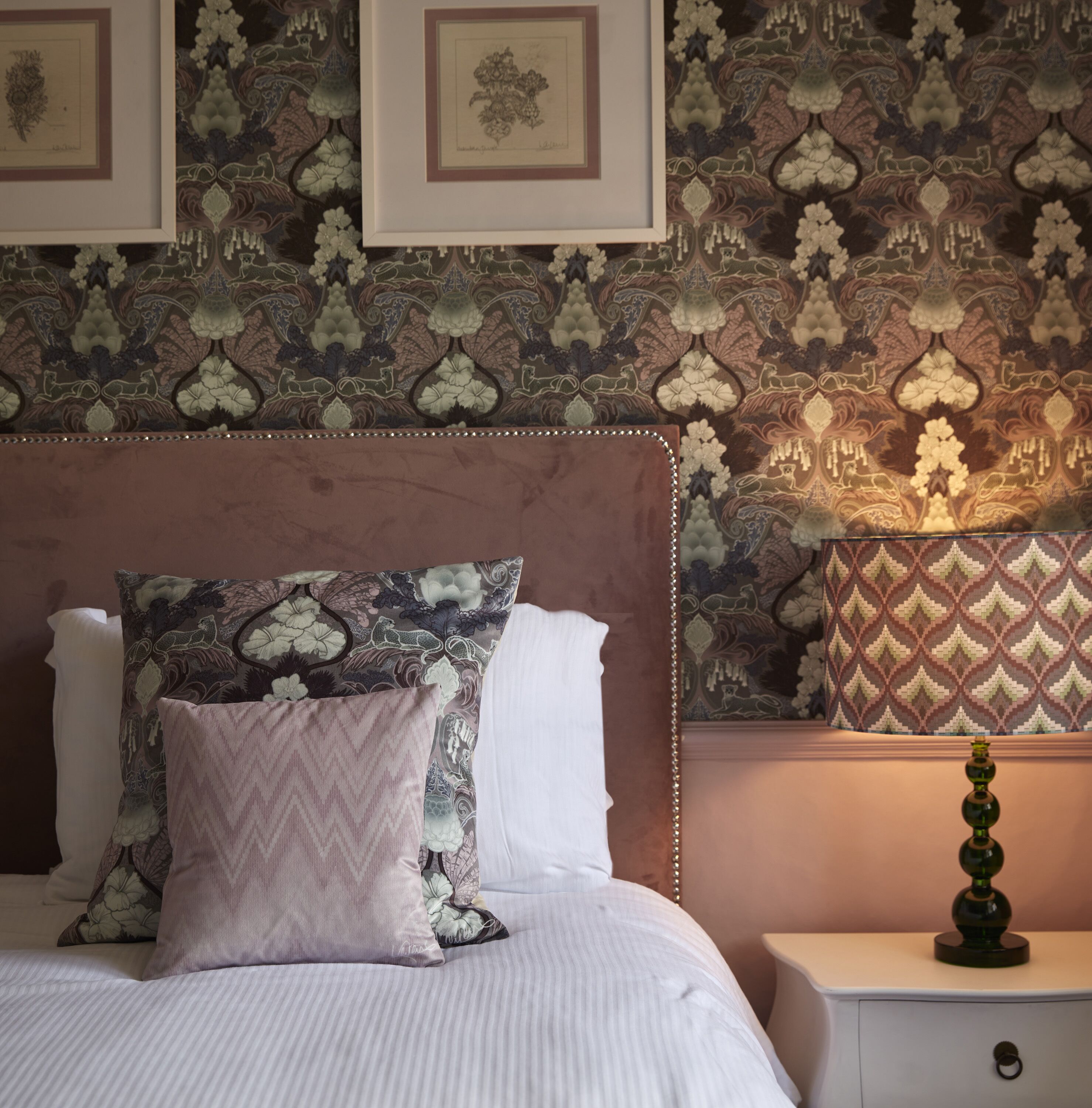 Une chambre à coucher avec un papier peint à motifs, un lit avec deux oreillers, une table de chevet avec une lampe et des œuvres d’art encadrées accrochées au mur.