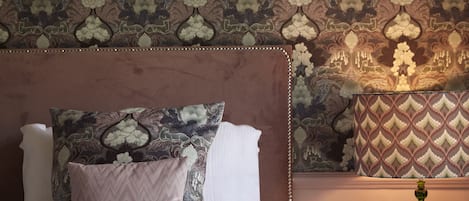 Une chambre à coucher avec un papier peint à motifs, un lit avec deux oreillers, une table de chevet avec une lampe et des œuvres d’art encadrées accrochées au mur.