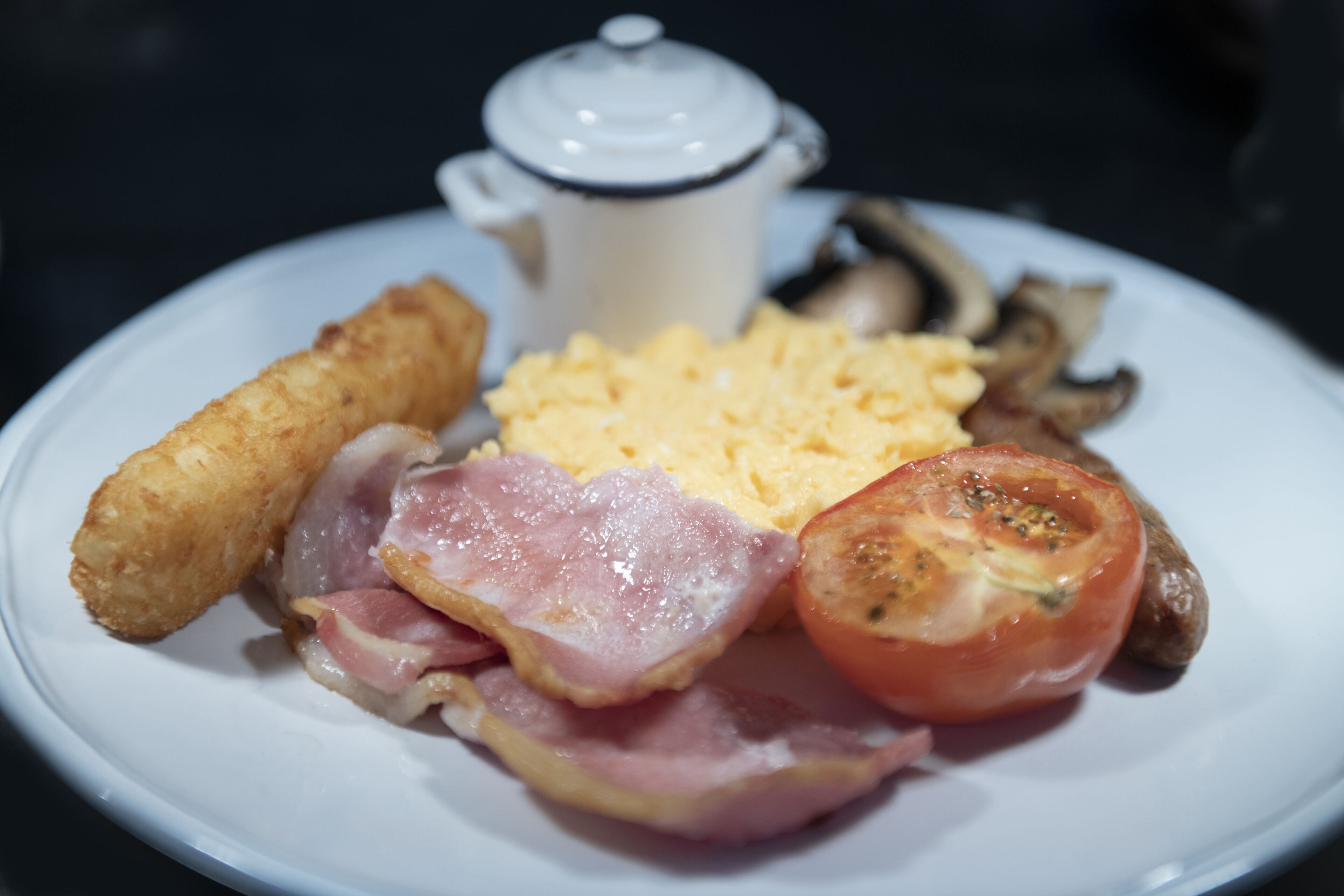 A plate with scrambled eggs, bacon, a fried item, a tomato, and a small pot of coffee.