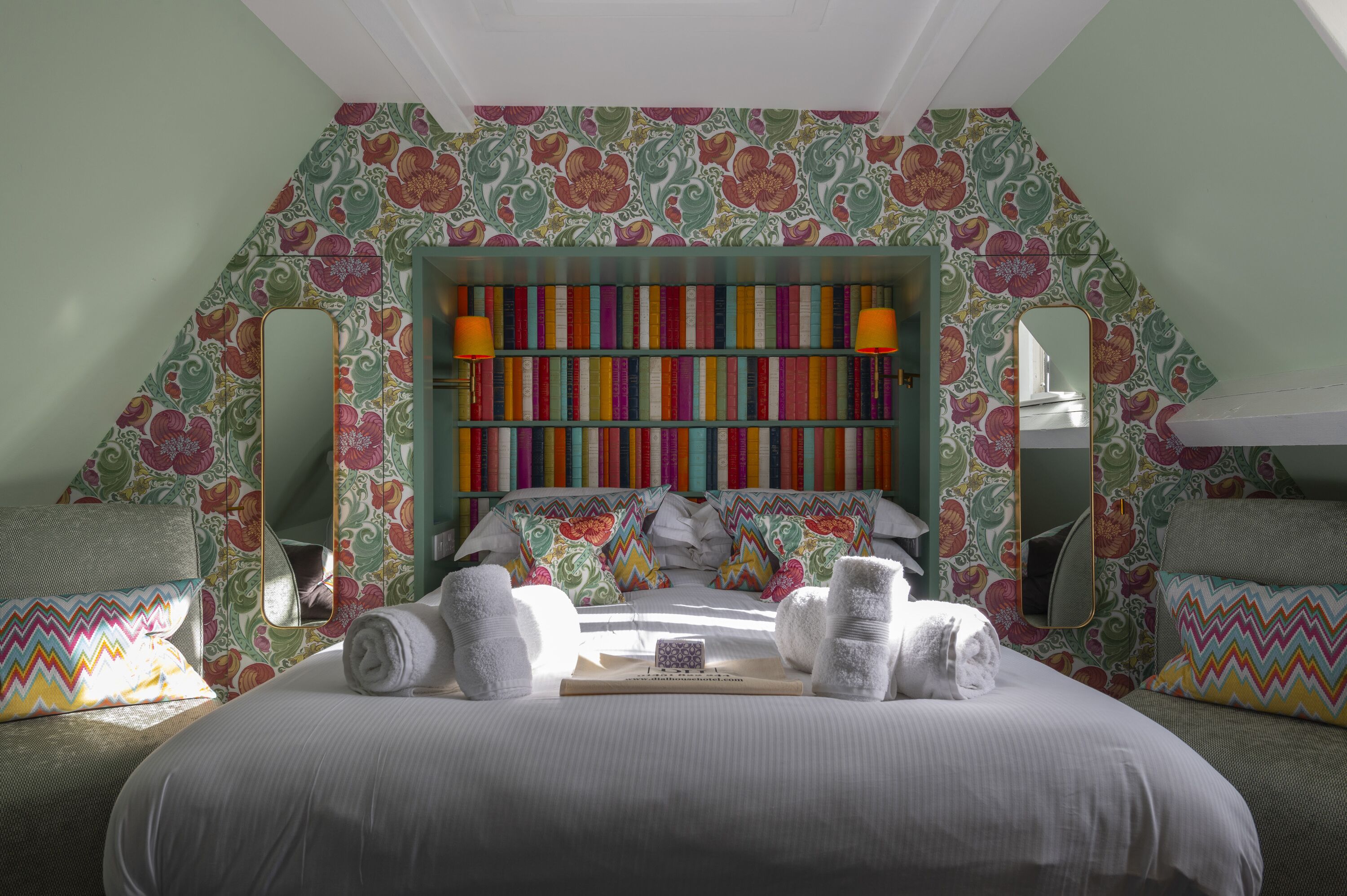 Un dormitorio con una estantería colorida, una cama con ropa de cama blanca y almohadas estampadas.
