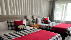 A hotel room with two beds, each with a checkered bedspread and red accents, a wooden nightstand with a plant, and a large window with curtains.