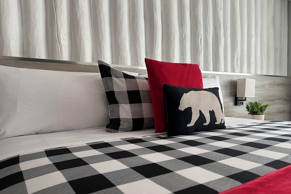 Una cama bien hecha con colcha a cuadros blancos y negros, dos almohadas con estampado a cuadros, una roja y otra con la silueta de un oso.
