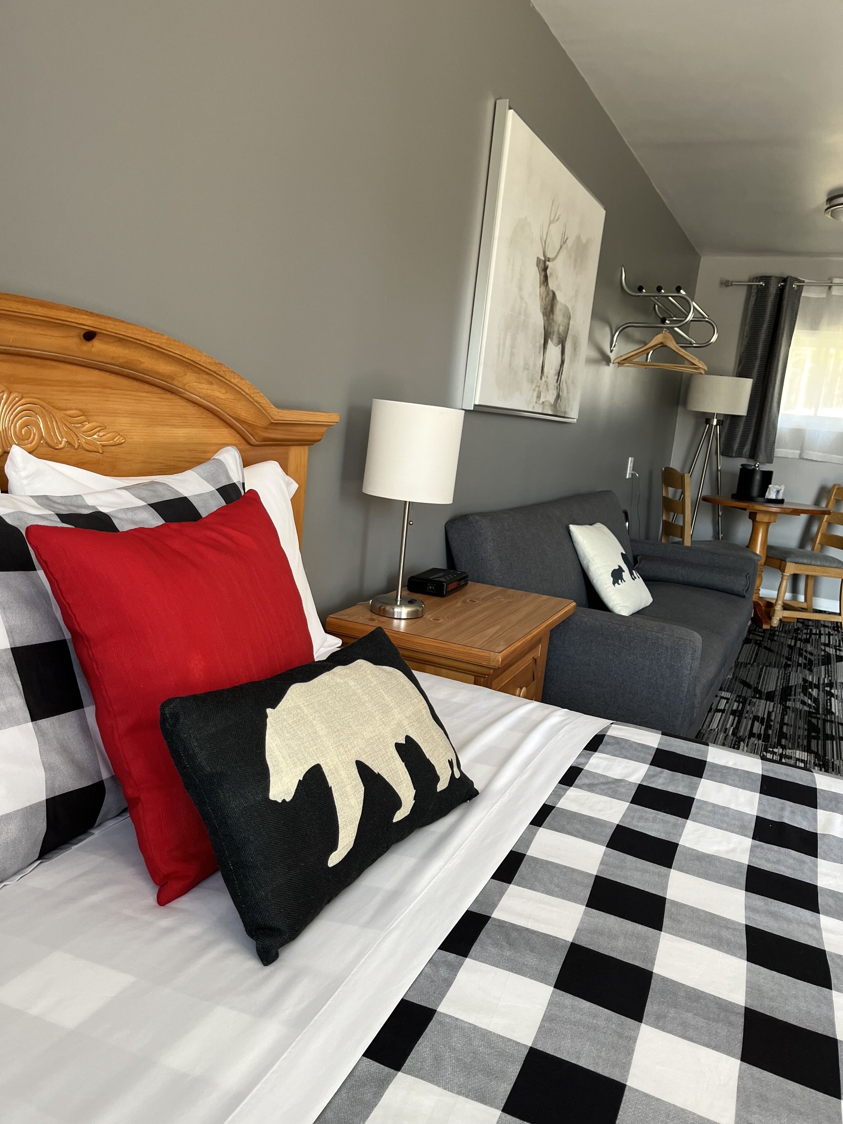 A hotel room with a large bed, a grey sofa, a wooden nightstand with a lamp, a table with chairs, and a painting on the wall.