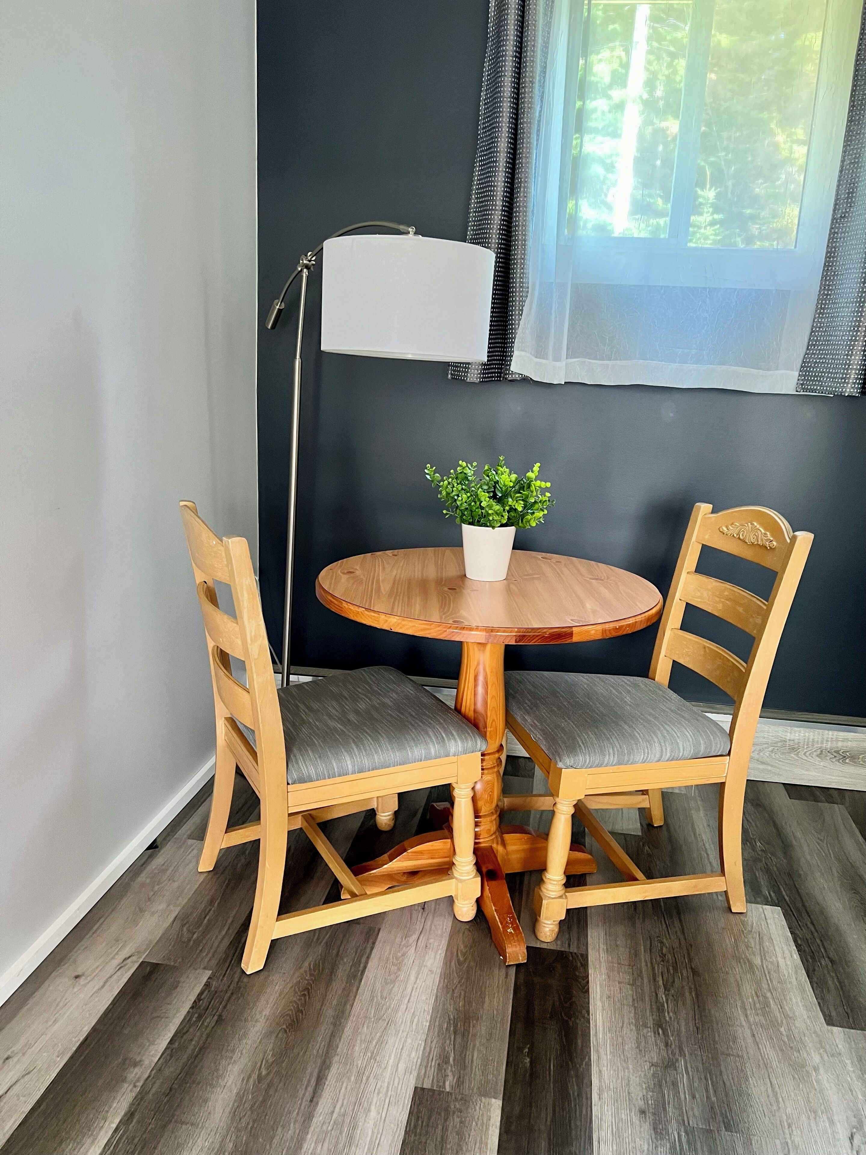 A dining area with a round wooden table, two wooden chairs with grey cushions, a white floor lamp, and a potted plant.