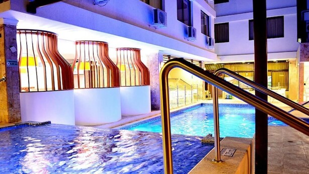 An indoor swimming pool with a glass floor, metal railings, and a view of the pool area through large windows with vertical blinds. - Hotel La Riviera (Santa Marta)