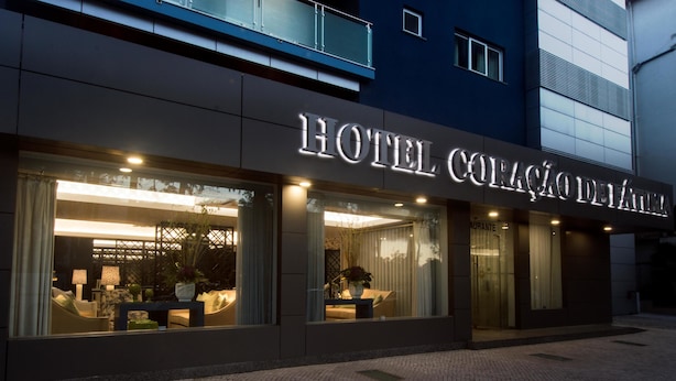 Exterior de un hotel con señalización iluminada, amplias ventanas y un área de entrada pavimentada.