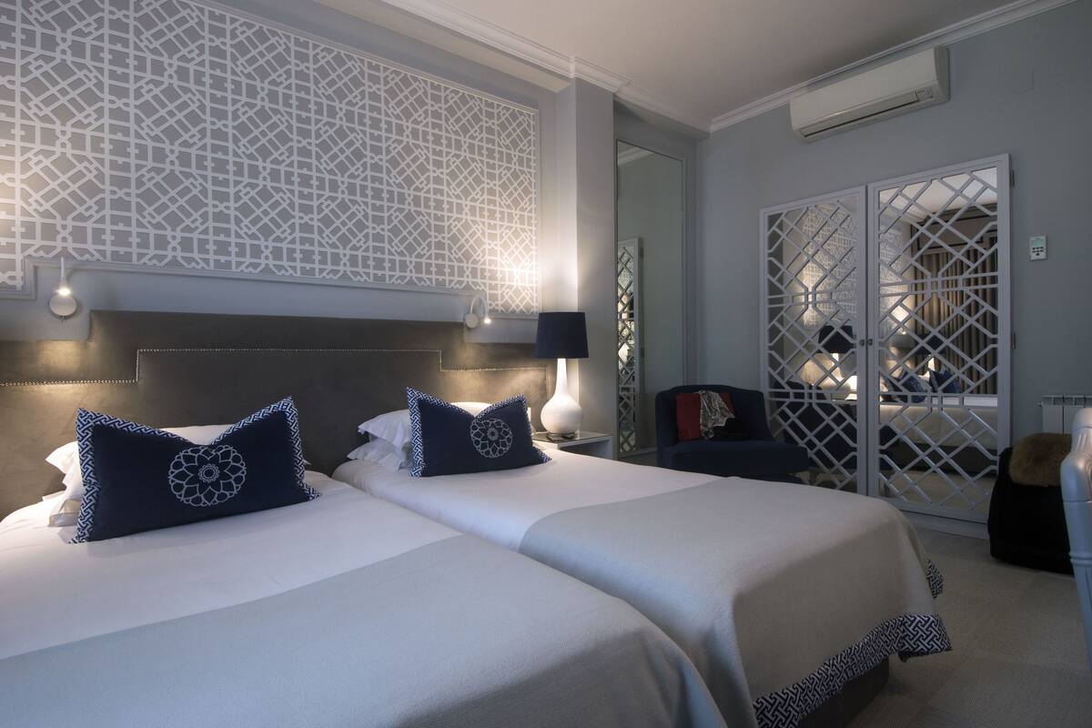Habitación de hotel con dos camas, un panel decorativo en la pared, una zona de estar con una silla y vistas a través de un ventanal.