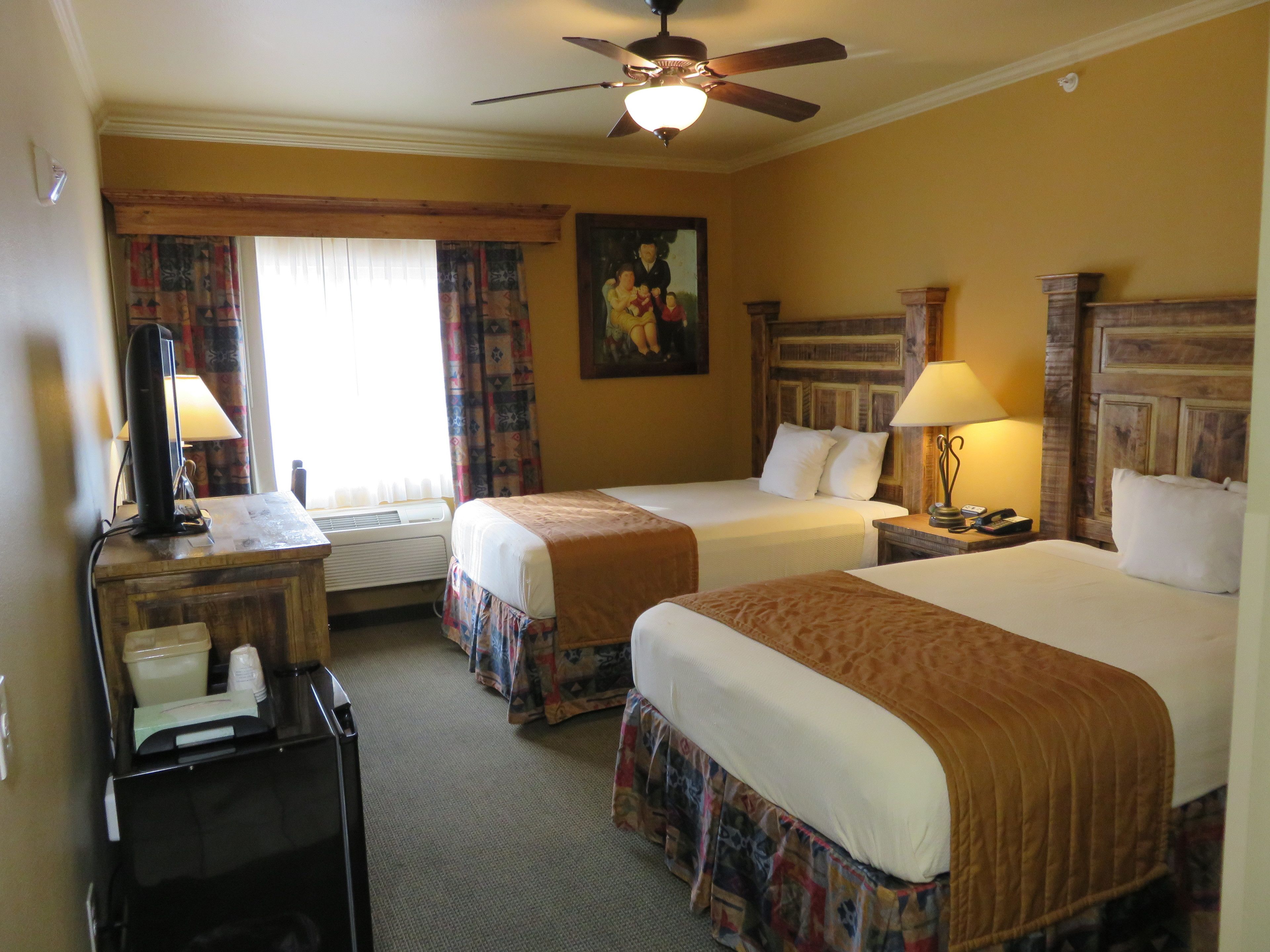 A hotel room with two beds, a desk with a lamp, a ceiling fan, and a painting on the wall.