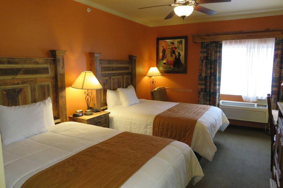 A hotel room with two beds, a ceiling fan, a painting on the wall, and an air conditioning unit.