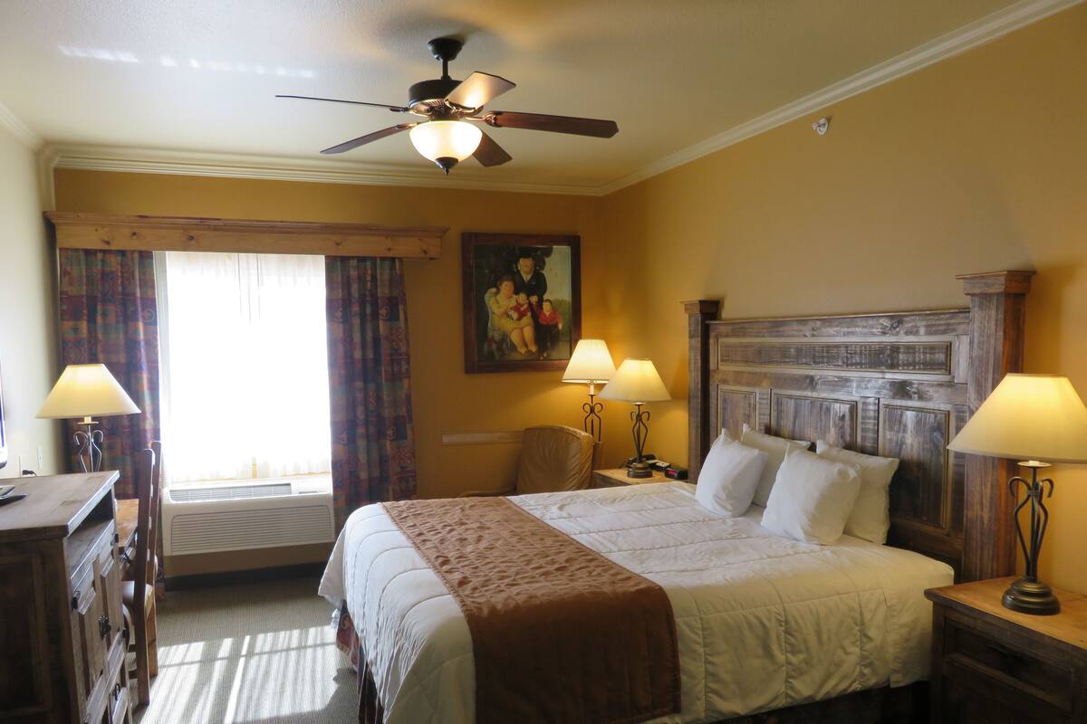 A hotel room with a bed, two bedside lamps, a ceiling fan, a painting on the wall, and a window with curtains.