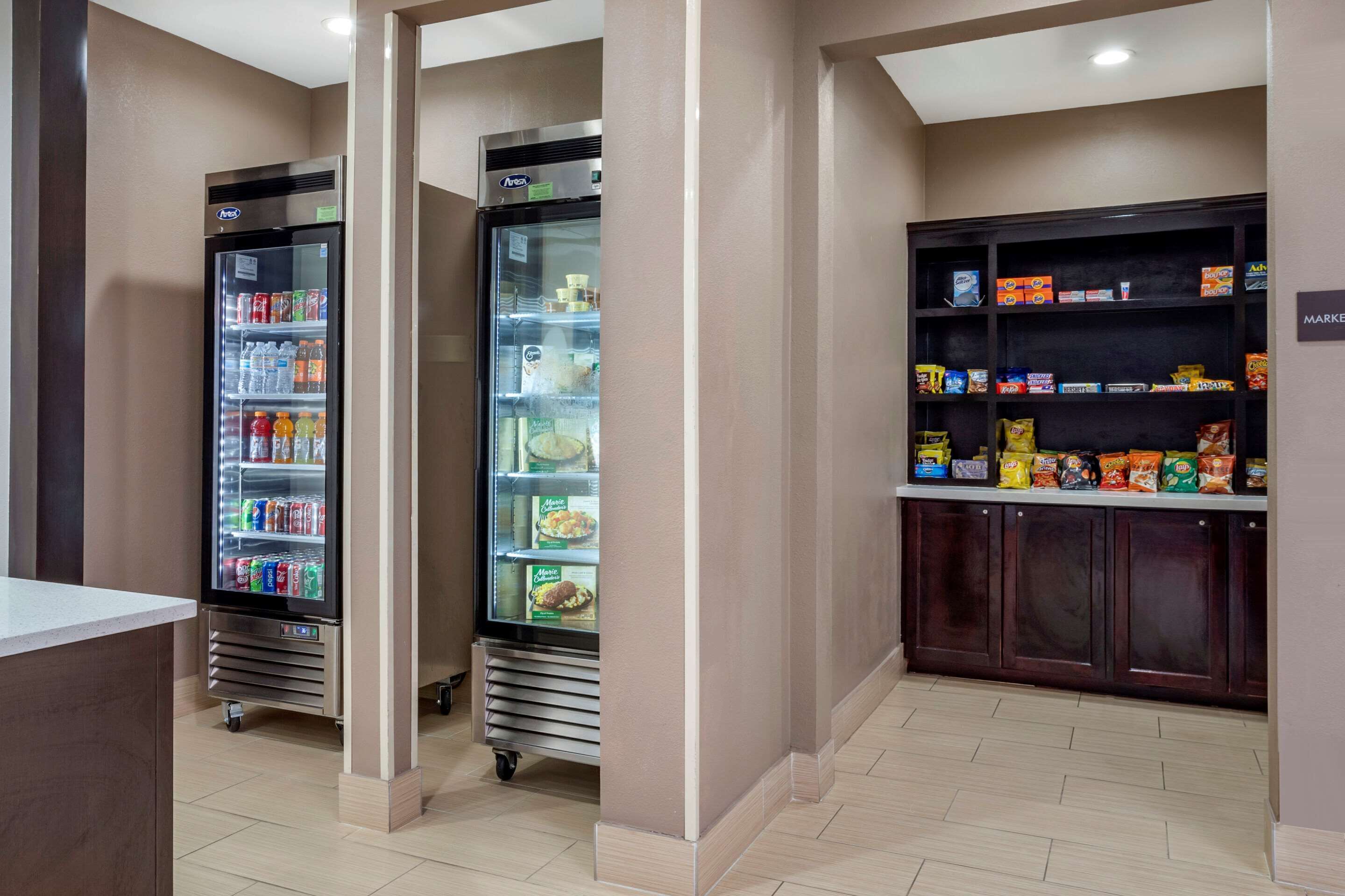 A hotel lobby with two refrigerated beverage dispensers, a snack bar with various packaged foods, and a reception desk.