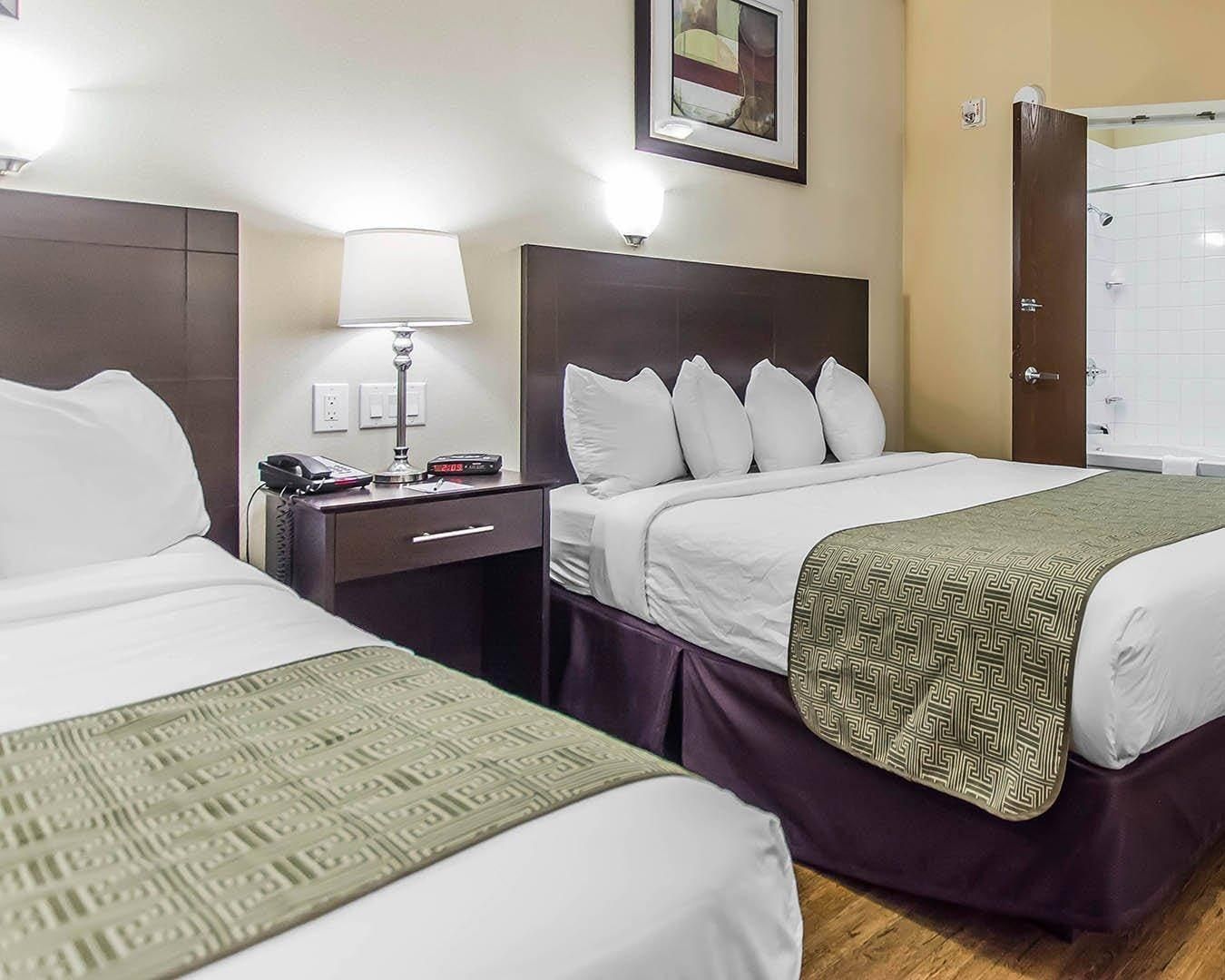A hotel room with two beds, a nightstand with a lamp, a clock, and a bathroom visible through an open door.