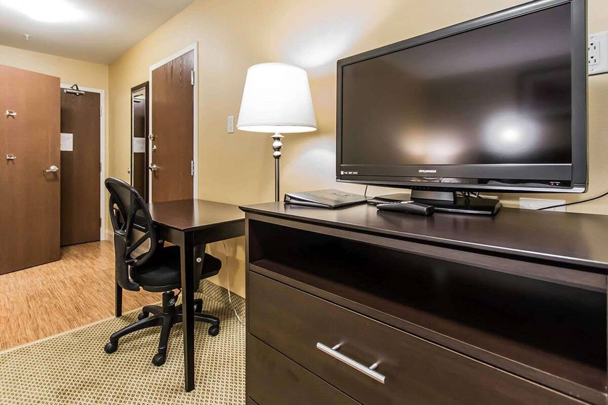 A hotel room with a dark wooden desk, a black office chair, a flat-screen TV, a lamp, and a filing cabinet.