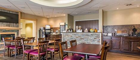 A restaurant with a central dining area featuring wooden tables and chairs, a stone accent wall, and a kitchen area with appliances and a coffee machine.
