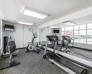 Un gimnasio con cintas de correr, elíptica y bicicleta estática. Hay relojes en la pared, un televisor y una ventana con vistas a edificios.