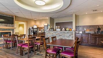 A restaurant with a central dining table surrounded by chairs, a kitchen area with appliances, and a stone accent wall.