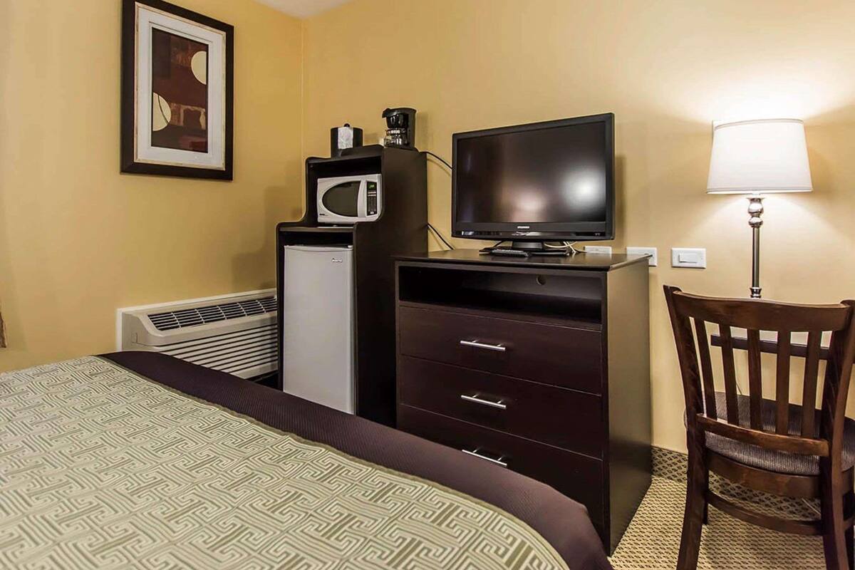 A hotel room with a bed, a television, a microwave, a refrigerator, a desk with a lamp, and a chair.