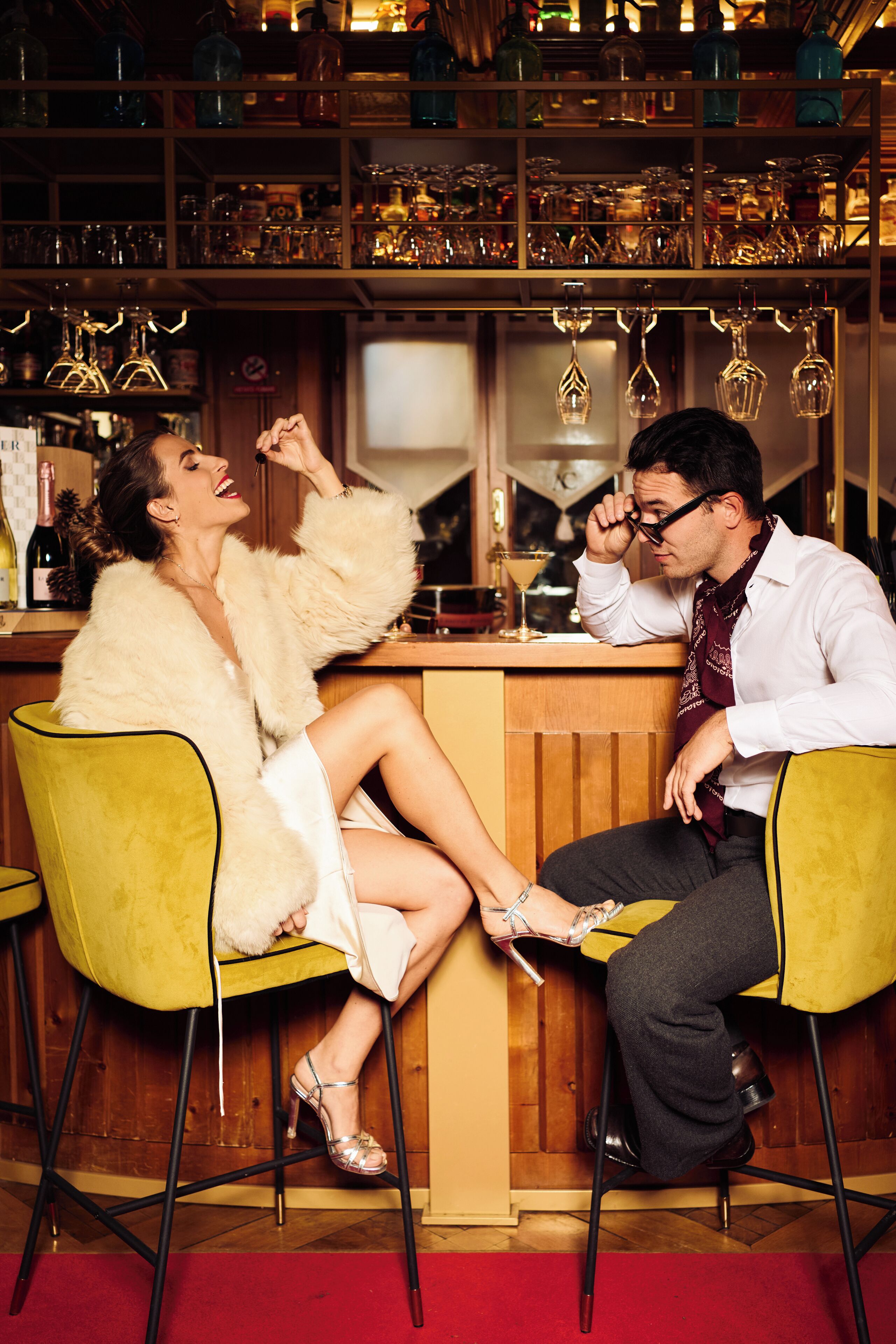 Zona de bar con dos personas sentadas en sillas amarillas, una barra de madera y estantes con cristalería.