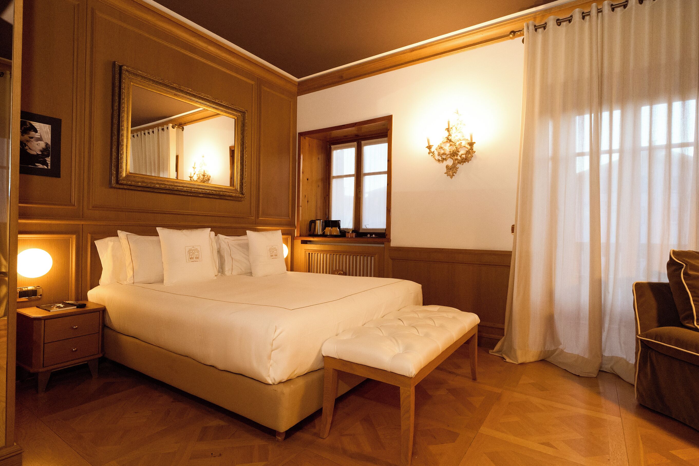 Habitación de hotel con una cama grande, una mesita de noche, un banco, una silla, un espejo, una ventana y un candelabro.