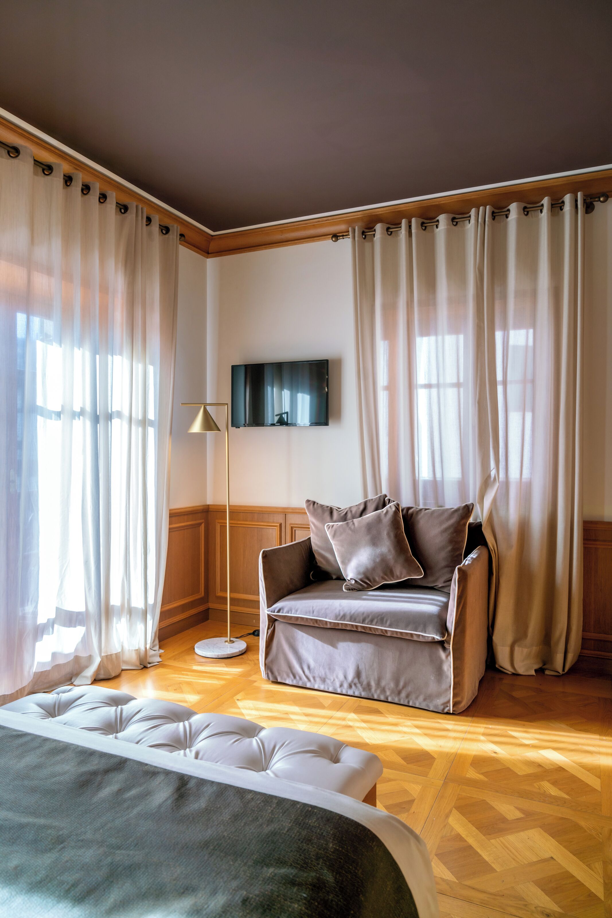 A hotel room with a wooden floor, a leather sofa with cushions, a flat-screen TV mounted on the wall, and a large window with sheer curtains.