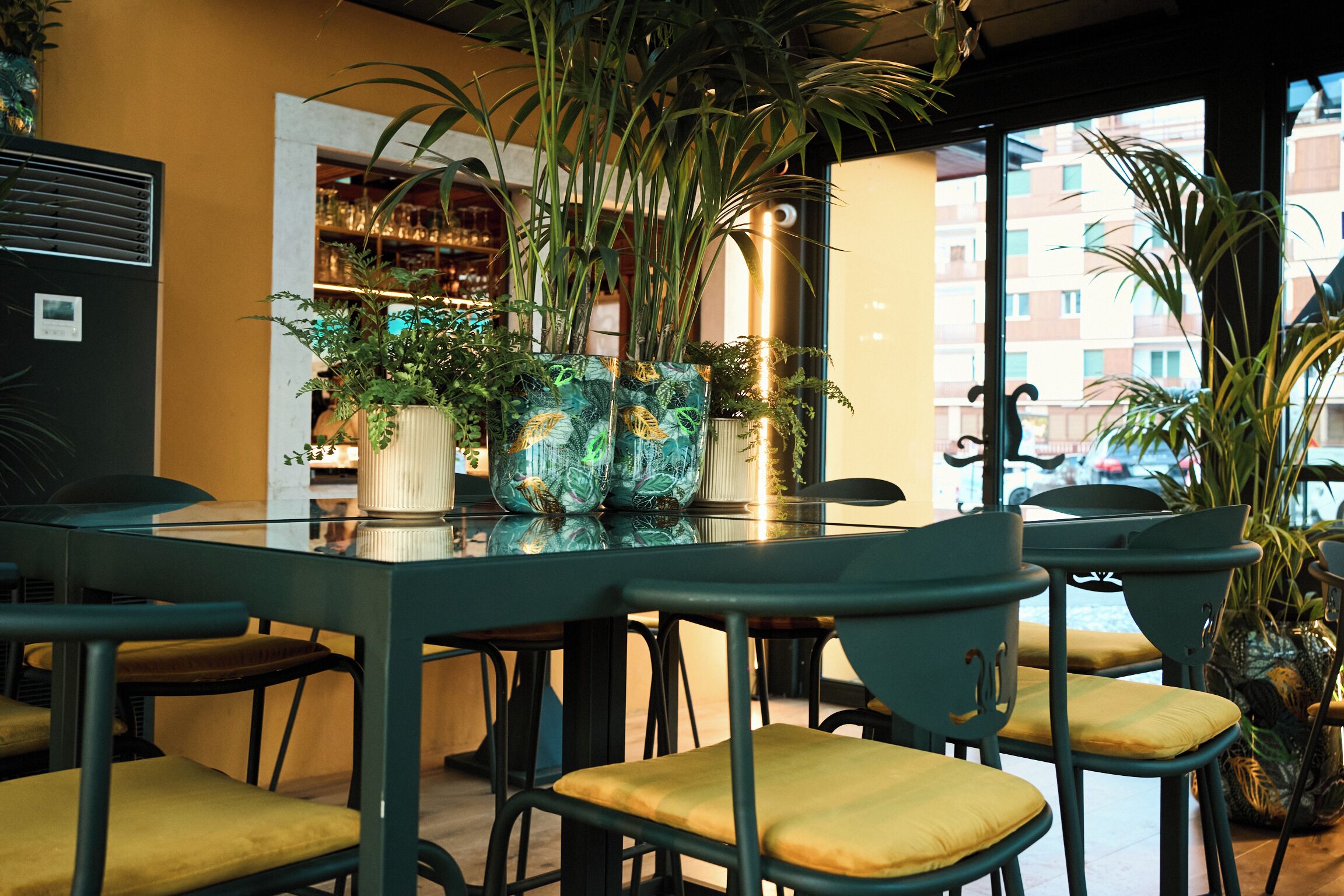 A restaurant with a glass table, green chairs, and potted plants.