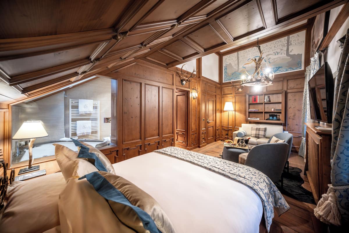 Une chambre spacieuse avec un grand lit, un coin salon comprenant un canapé et des fauteuils, un lambris en bois et un lustre.