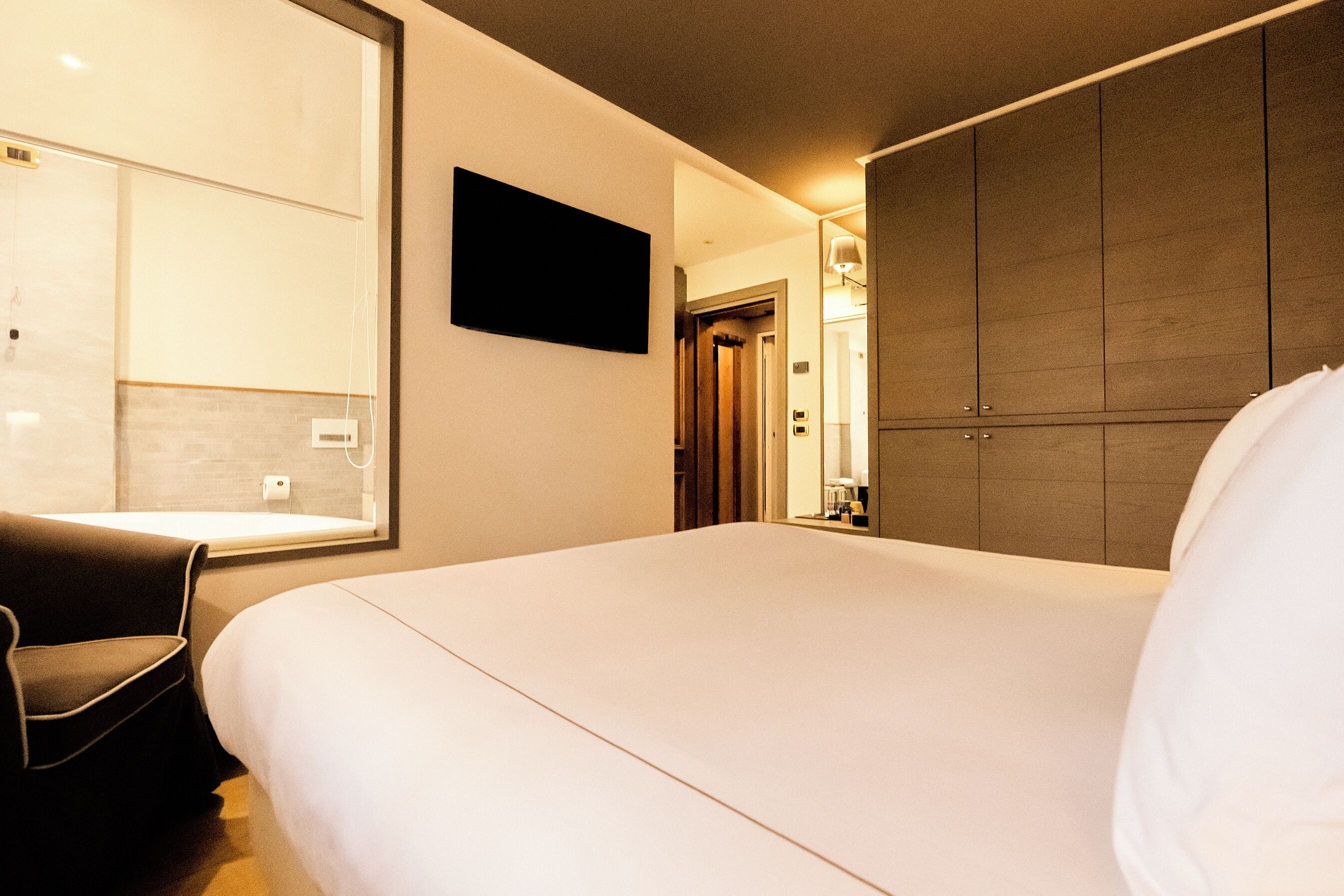 A hotel room with a large bed, a chair, a TV mounted on the wall, and a wooden wardrobe.