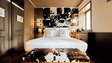Une chambre d’hôtel avec un grand lit, un banc en bois, un mur décoré d’un motif représentant une vache et une fenêtre.