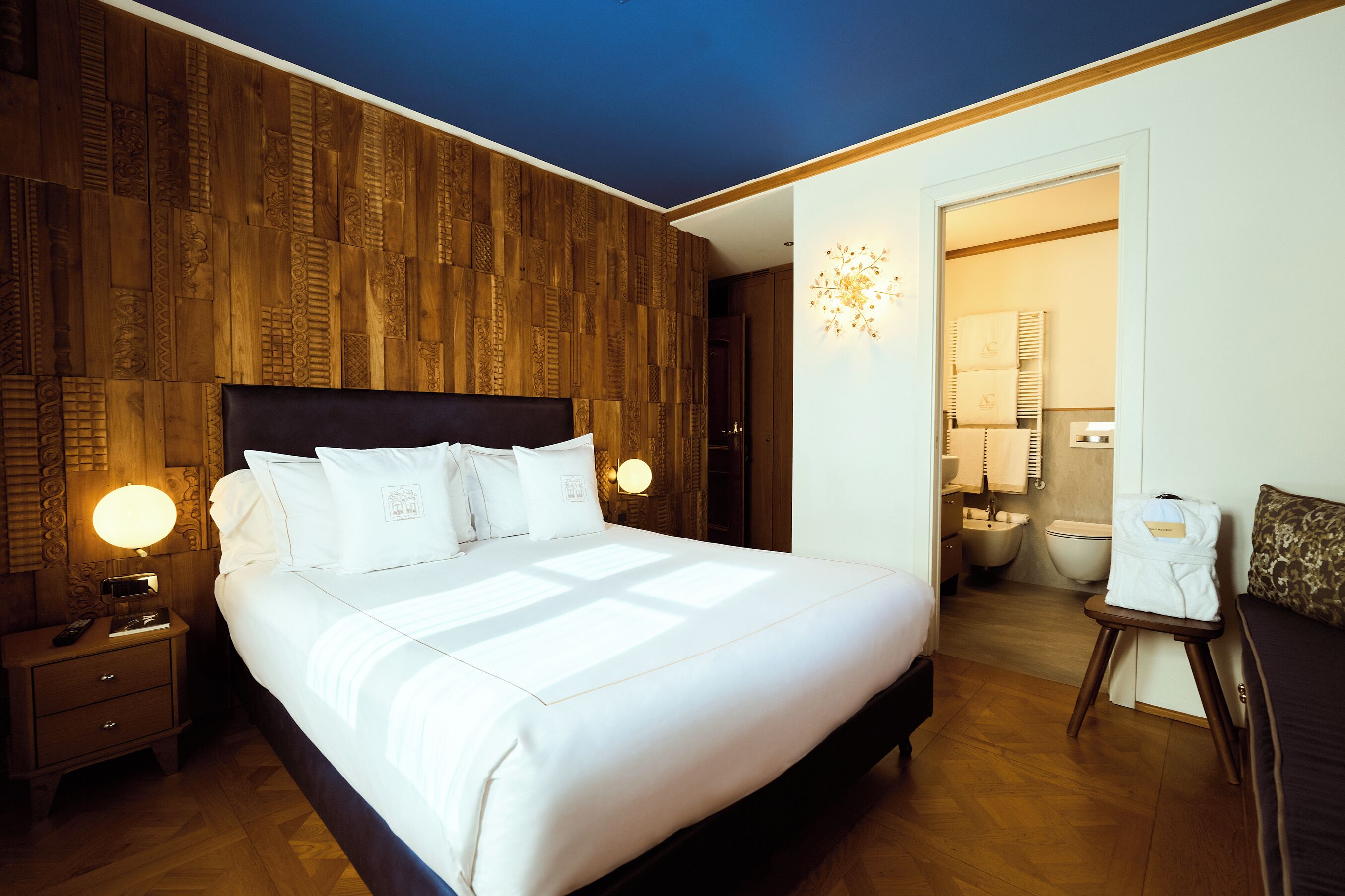 A hotel room with a large bed, wooden panel wall, bedside lamps, a small table with a robe, and a bathroom visible through an open door.