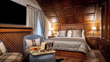 Habitación de hotel con techo de madera, cama grande con colcha estampada, zona de estar con bandeja de frutas y bebidas, y dos lámparas de noche.