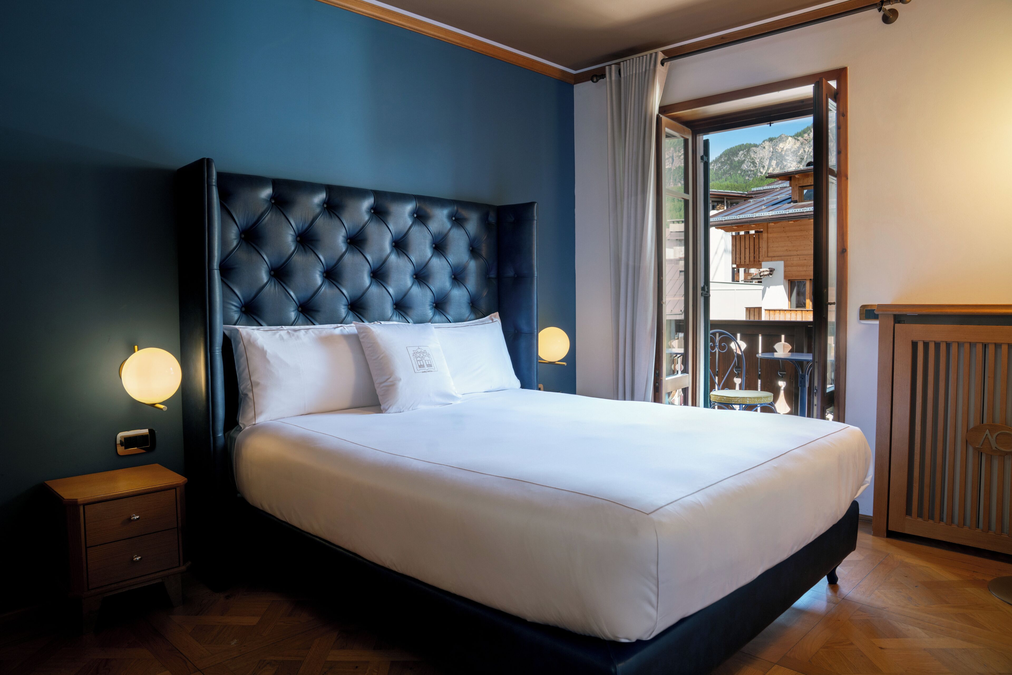 Un dormitorio con cabecera capitonada, mesita de noche de madera, cama con ropa blanca y vista a la montaña a través de un vano.