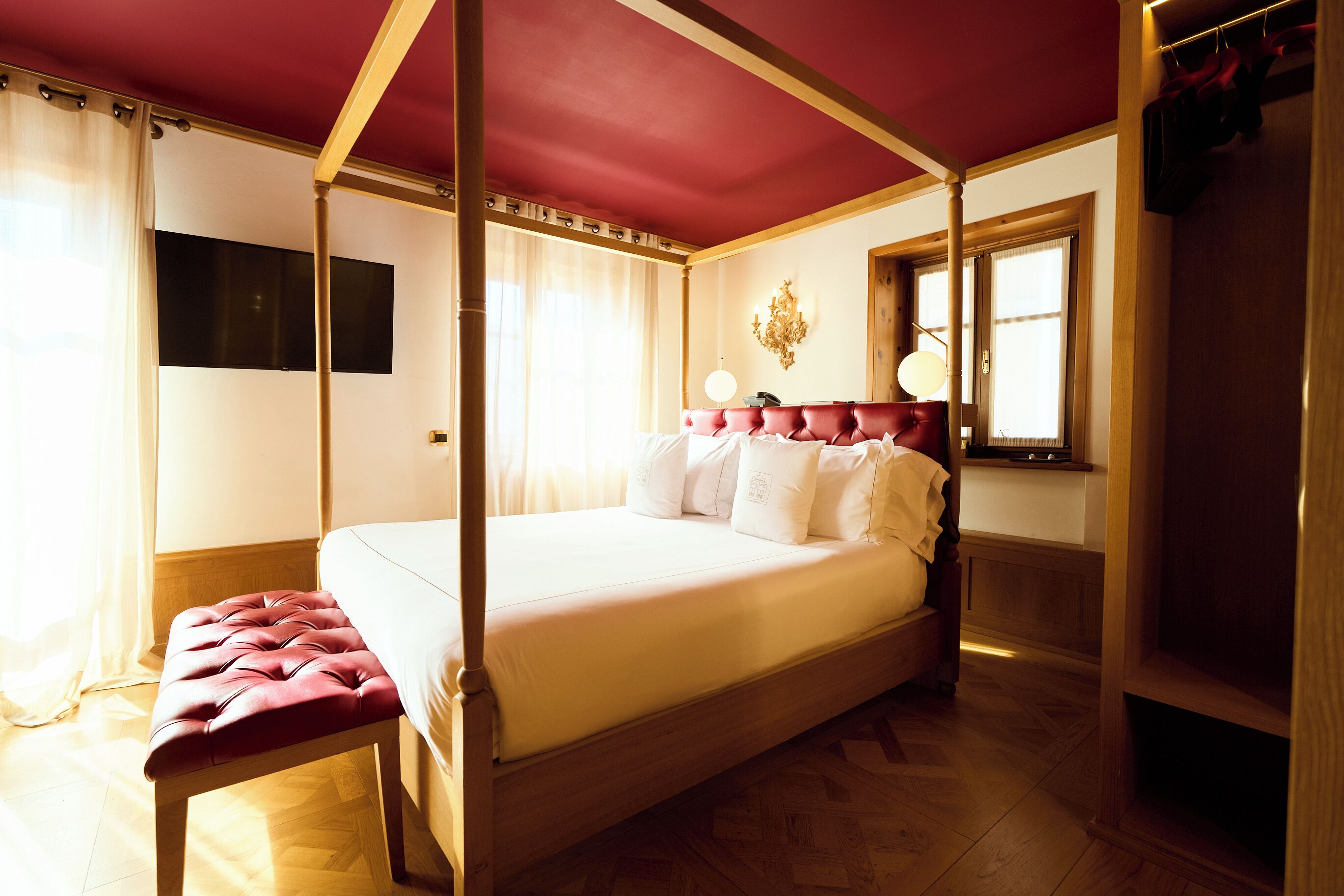 A four-poster bed with a canopy, a bench at the foot of the bed, a flat-screen TV mounted on the wall, and a window with curtains.