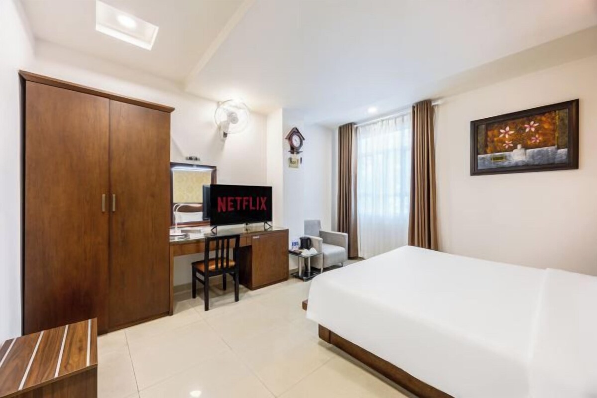 Ein Hotelzimmer mit einem Bett, einem Schreibtisch mit Computer und einem Fernseher, der 'NETFLIX' zeigt, einem Kleiderschrank, einem Sessel und einem Bild an der Wand.
