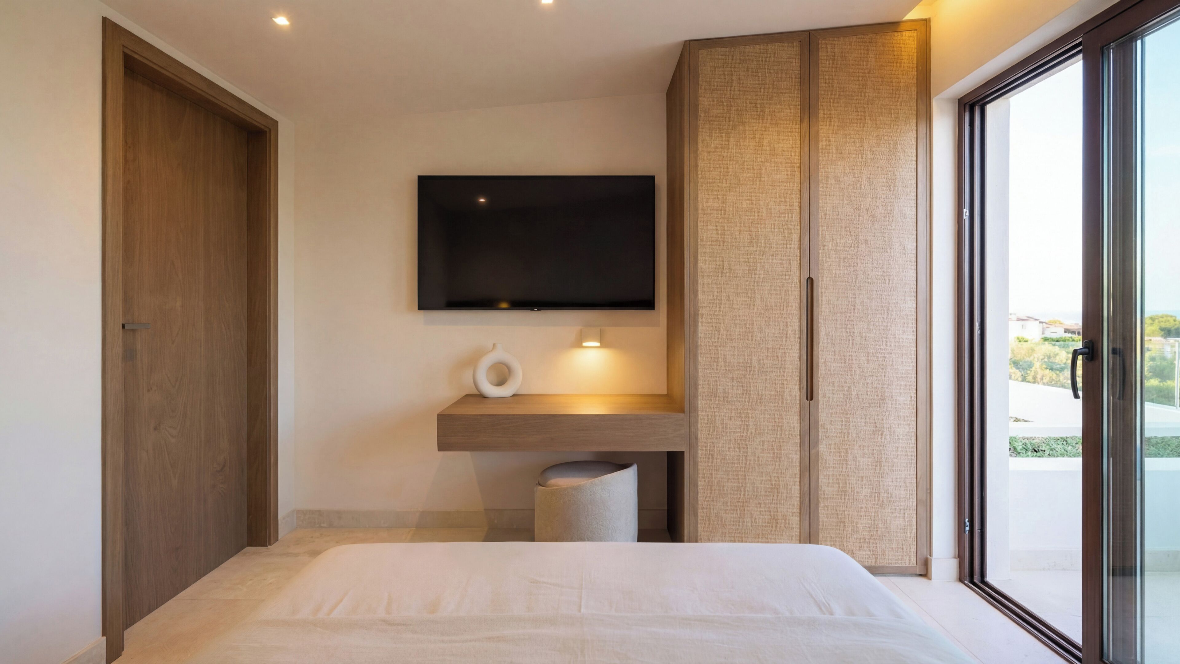 Un dormitorio moderno con una cama grande, un televisor montado, una mesita de noche pequeña y un ventanal corredero al exterior.