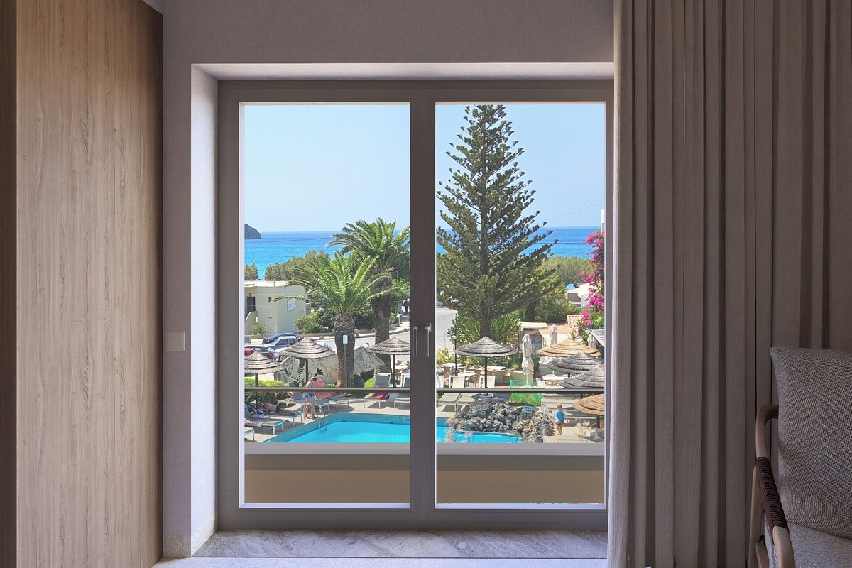 A room with a large sliding glass door leading to a pool area with a view of the sea and palm trees.
