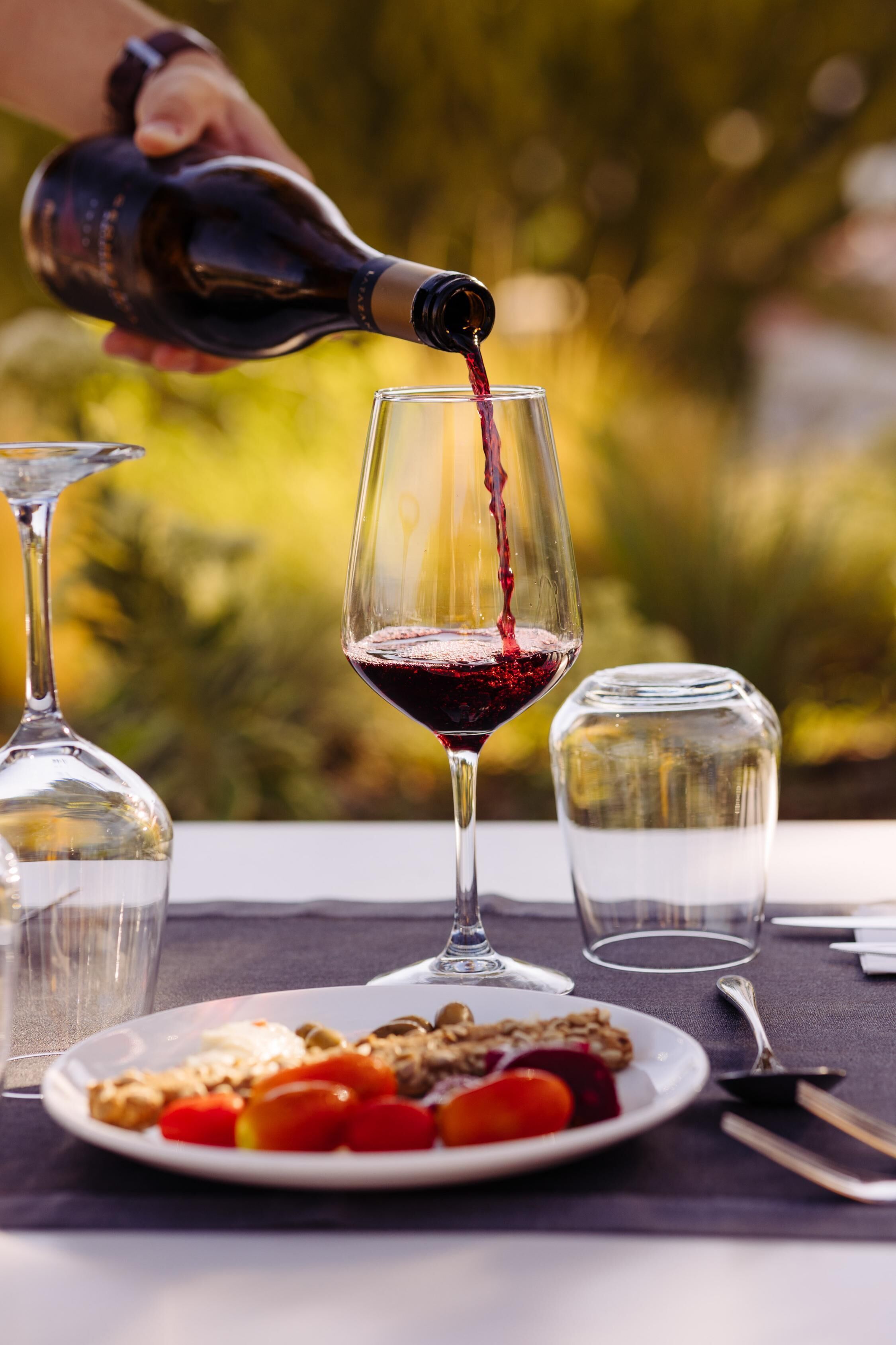 Une bouteille de vin rouge est versée dans un verre, et une assiette de nourriture ainsi qu'un verre d'eau sont posés sur une table.