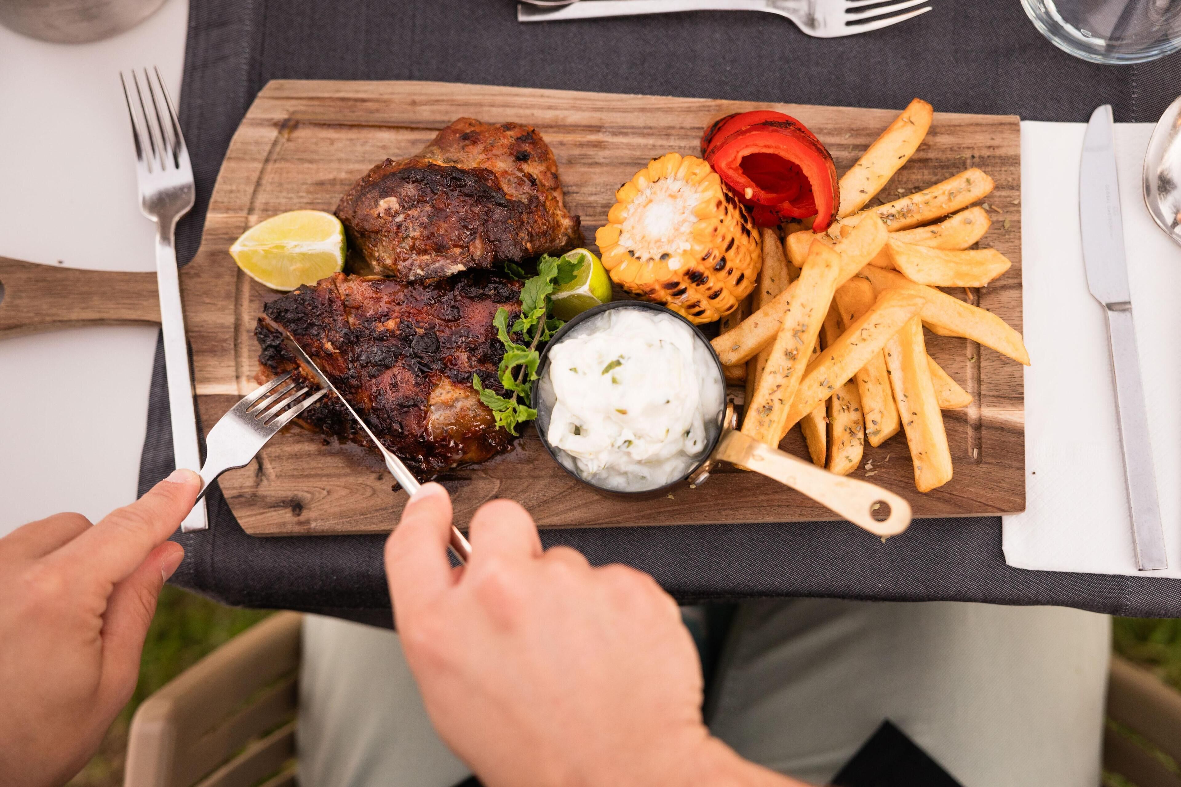 A wooden cutting board with grilled meat, French fries, a corn on the cob, and a small bowl of sauce.