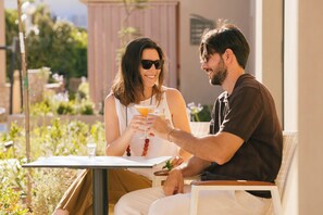 Dos personas sentadas en una mesa al aire libre, con bebidas, rodeadas de follaje.