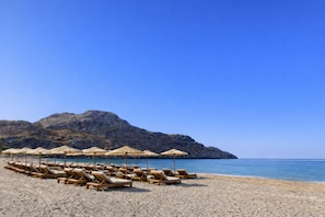 A beach with sun loungers and umbrellas, a clear sky, and a rocky mountain in the background.