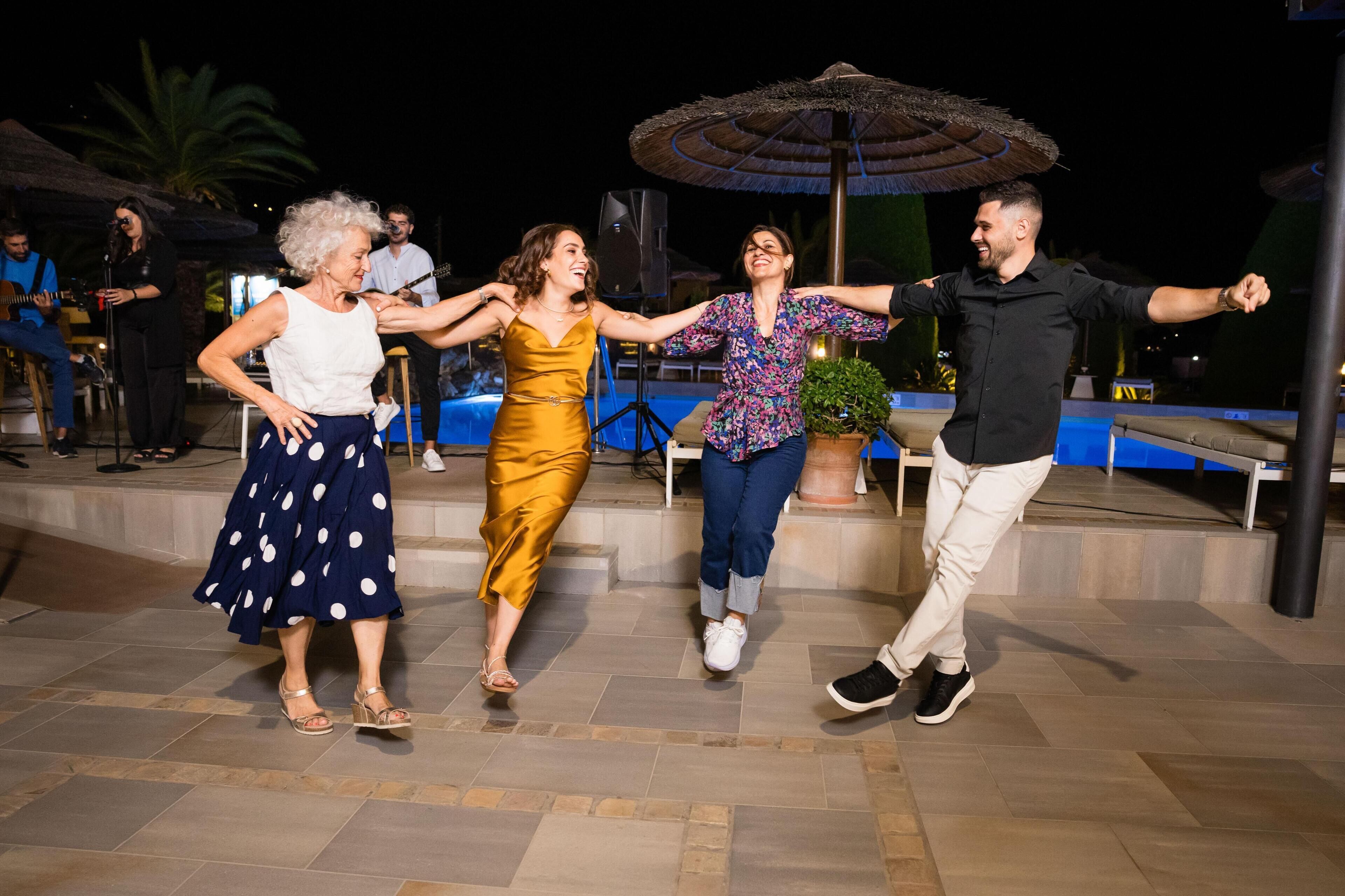 Un grupo de personas bailando en un espacio al aire libre con baldosas, con una piscina y sombrillas de fondo.