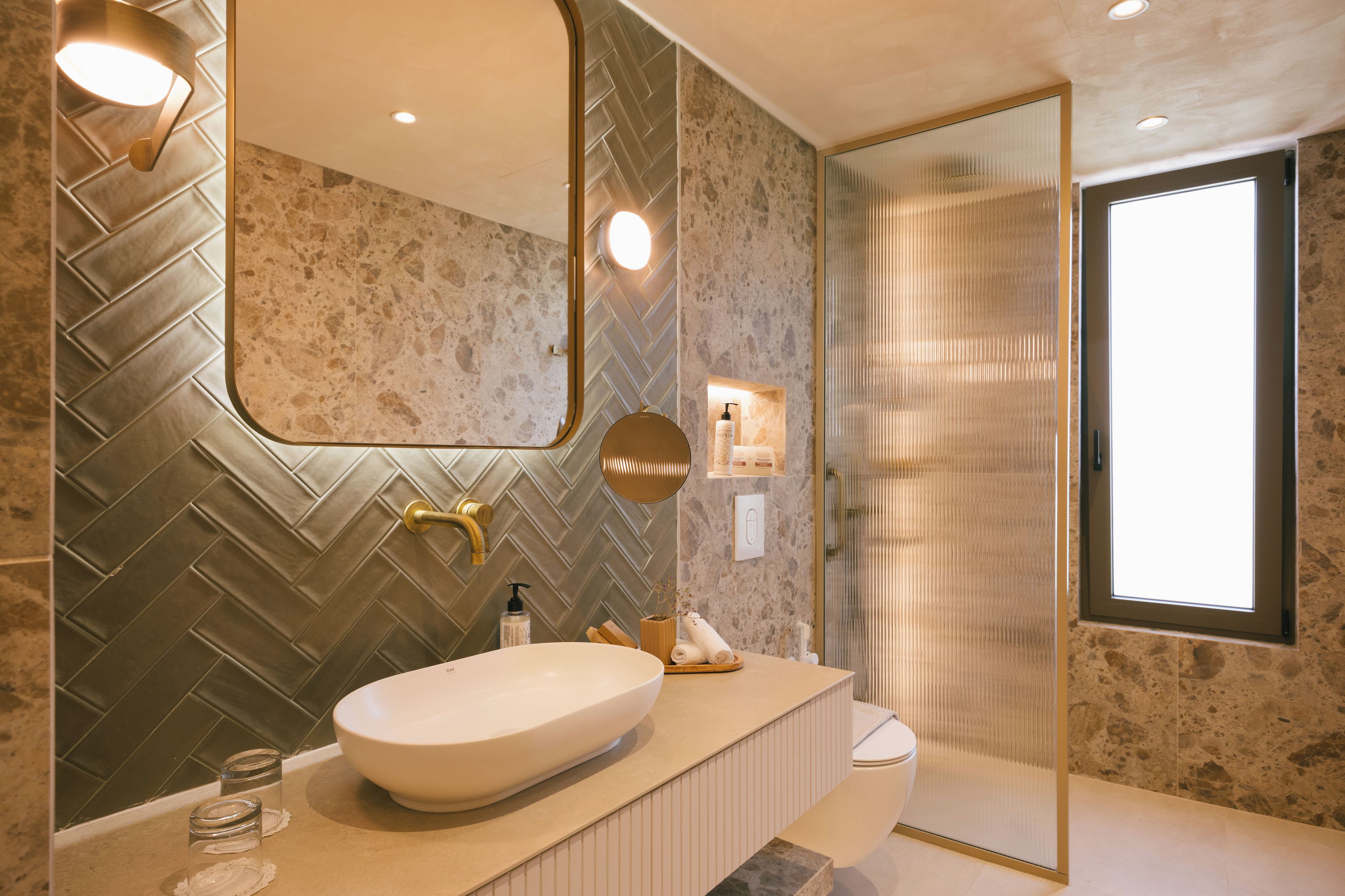 A modern bathroom with a herringbone-patterned wall, a large mirror, a freestanding sink, and a glass shower enclosure.