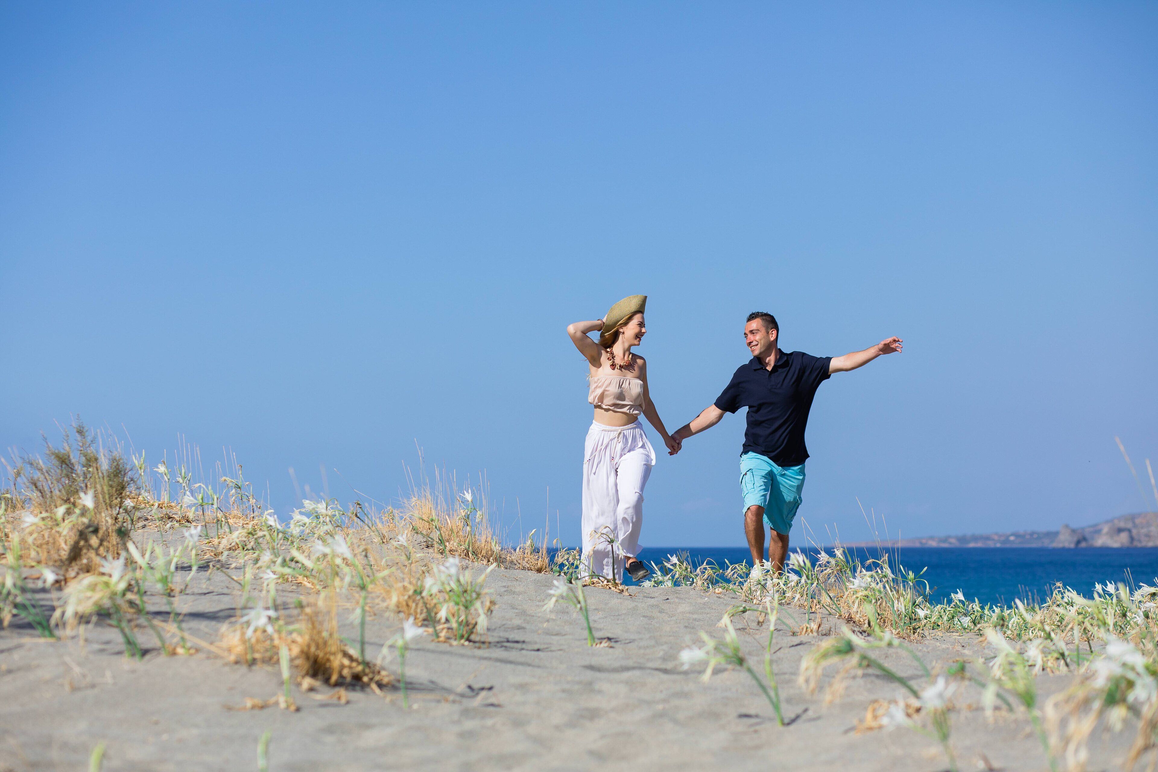 Una pareja camina de la mano por una playa arenosa con poca vegetación y un cielo azul despejado.