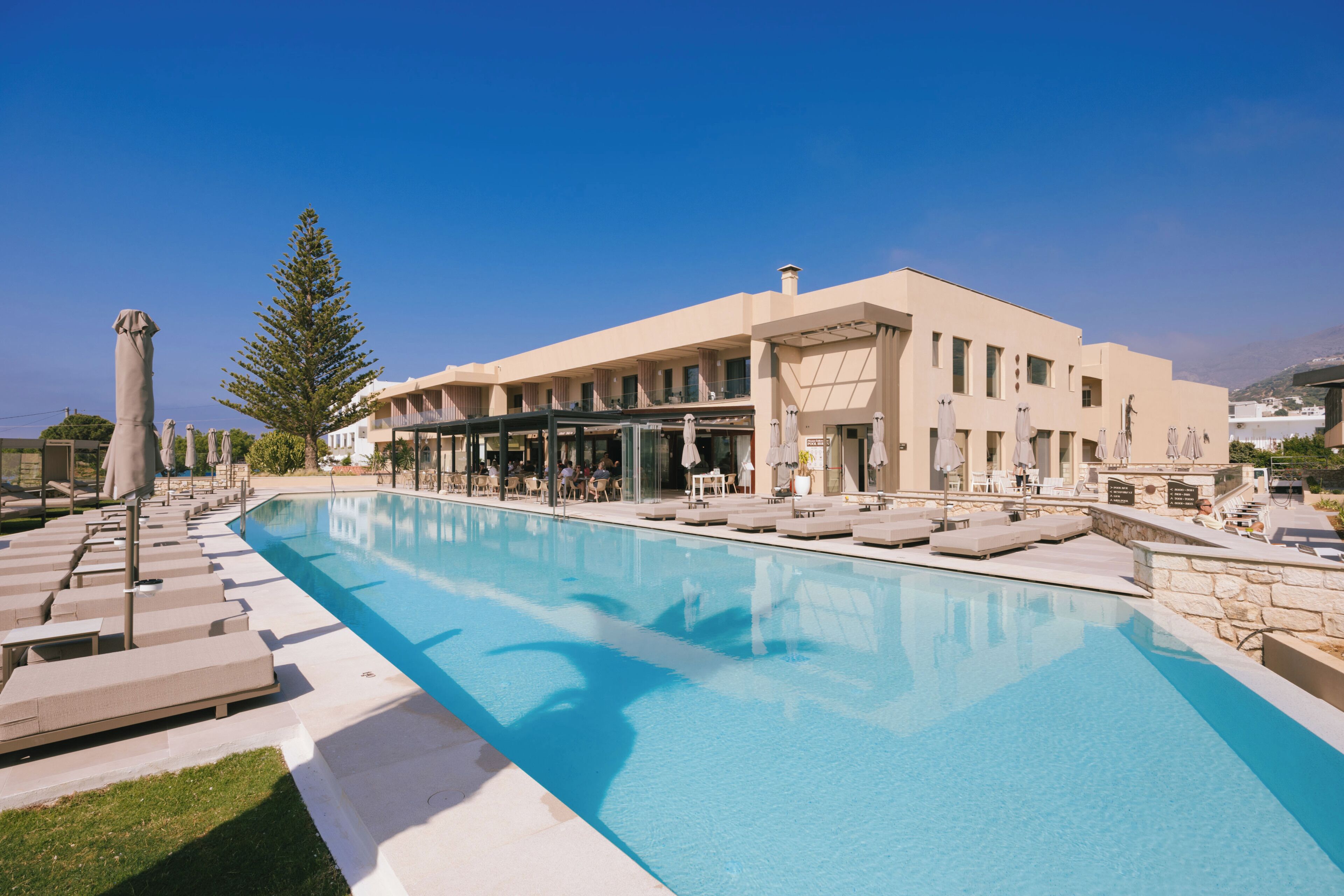 A poolside area with lounge chairs and umbrellas, a clear sky, and a building with multiple windows and balconies.