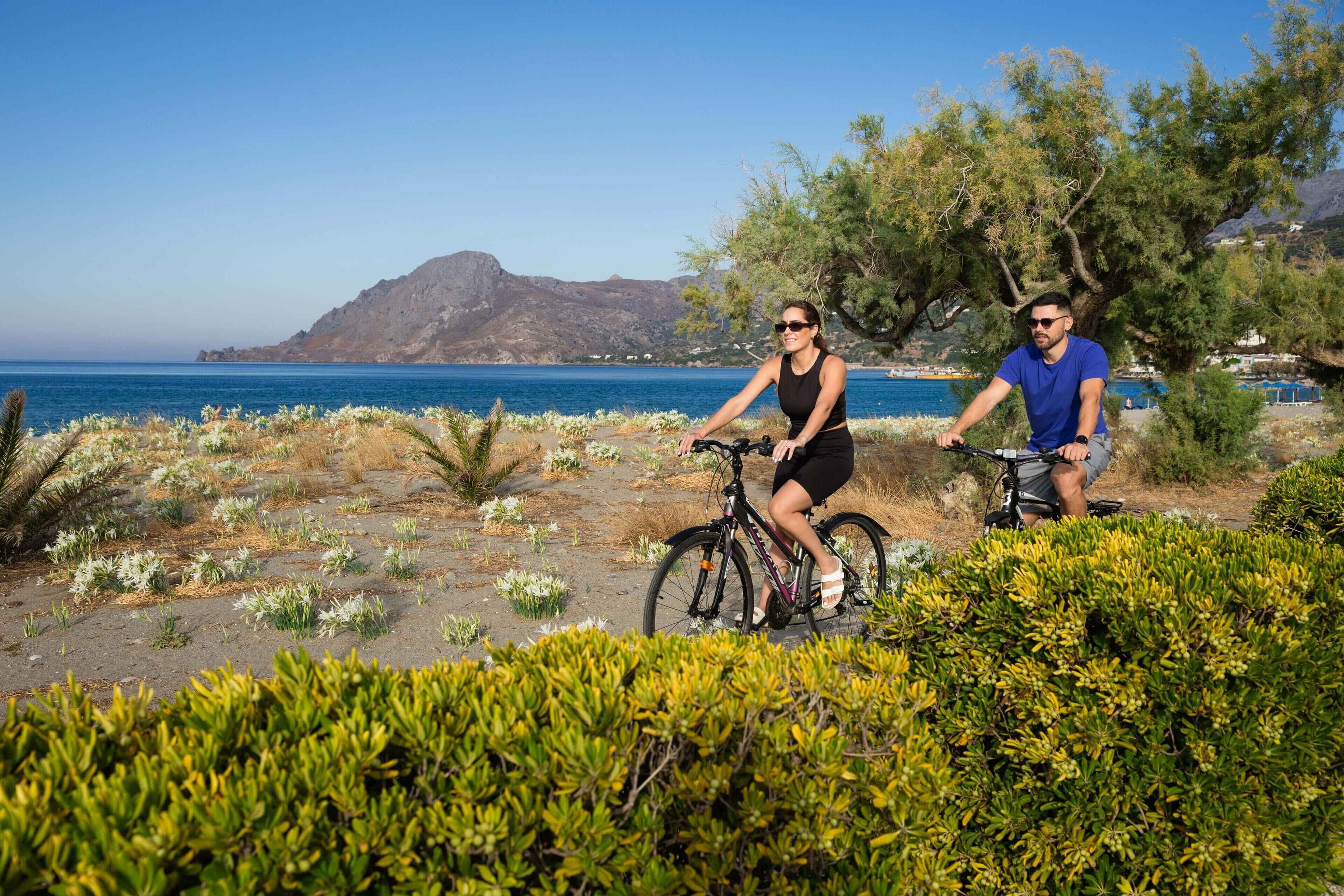 Two people on bicycles near a beach with a mountain in the background.
