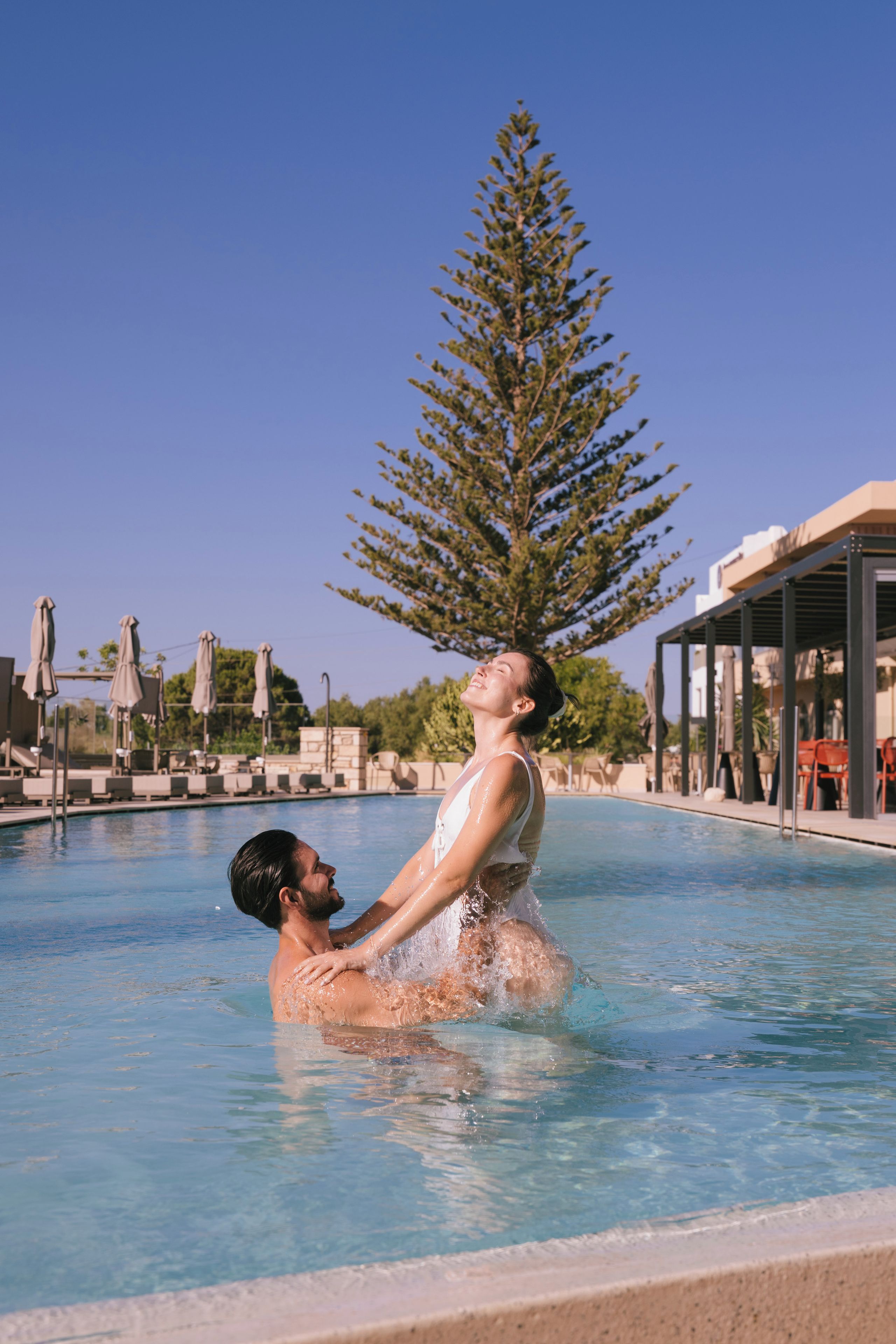 Una pareja en una piscina, con un cielo despejado y un árbol alto al fondo.