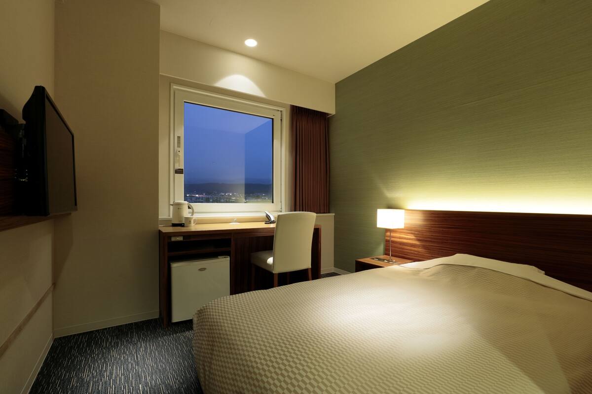A hotel room with a bed, a desk with a chair, a small fridge, a window with a view, and a nightstand with a lamp.