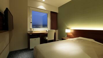 Ein Hotelzimmer mit Bett, Schreibtisch mit Stuhl, kleinem Kühlschrank, Fenster mit Blick und Nachttisch mit Lampe.